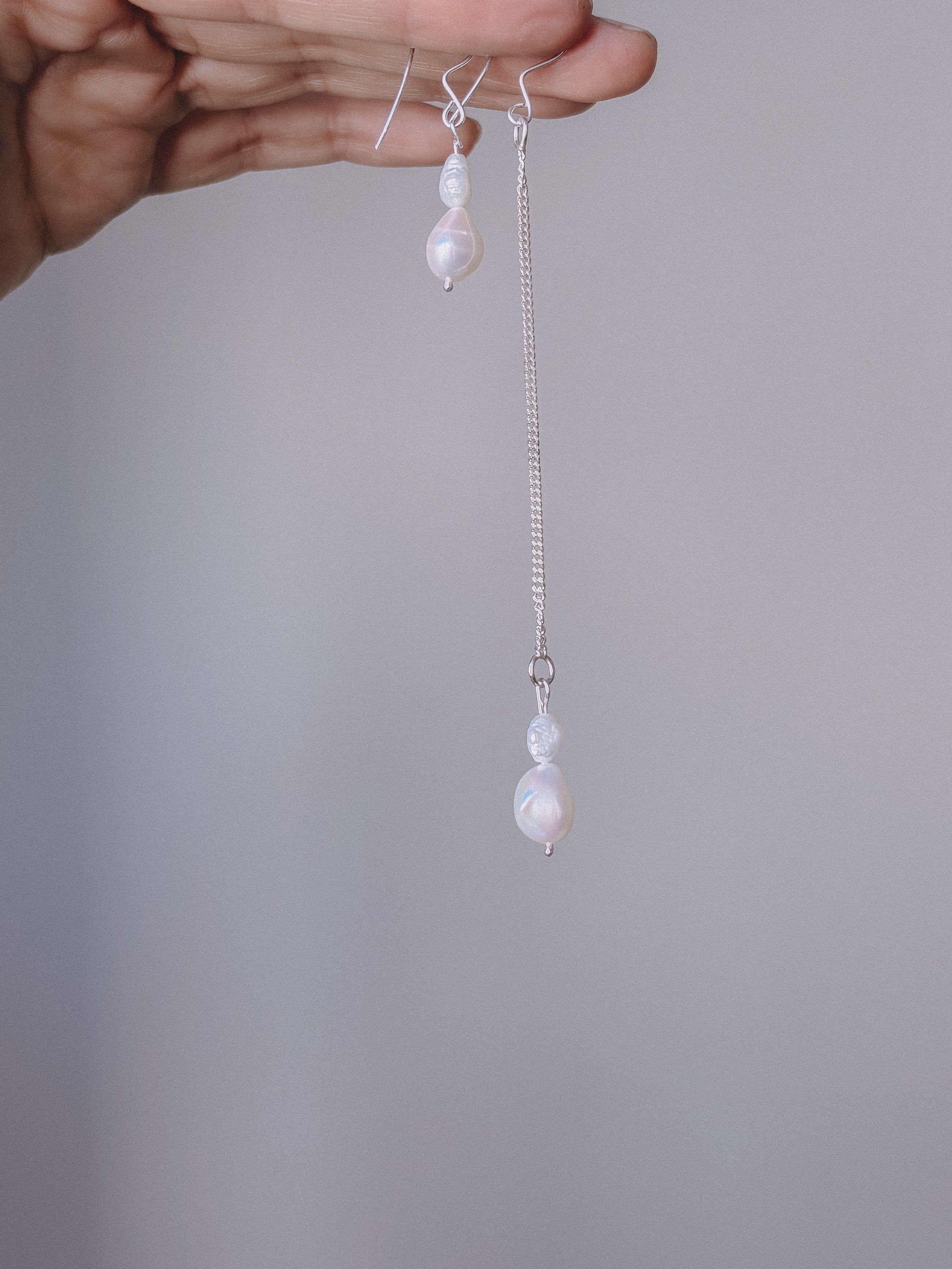 Pearl earrings with silver chains held by a hand against a gray background